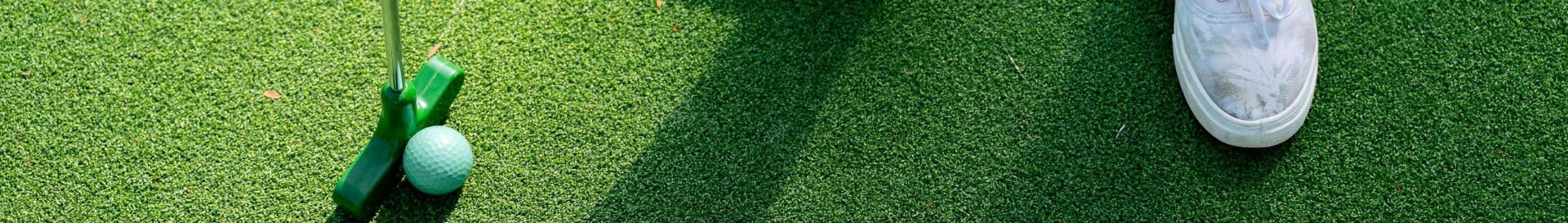 A person in white sneakers standing with a putter and golf ball on a green putting surface.